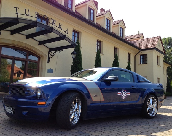 Blue sports car on cobblestone drive outside elegant cream boutique hotel with arched glass entrance and tiled roof