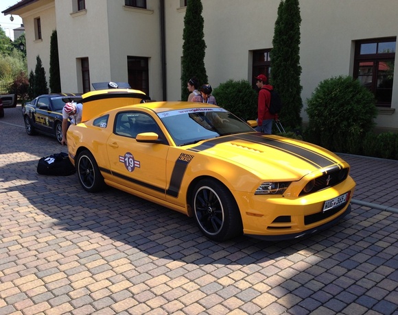 Yellow Ford Mustang with black racing stripes and rally decal parked on cobblestone driveway by hotel facade; trunk open, people loading luggage