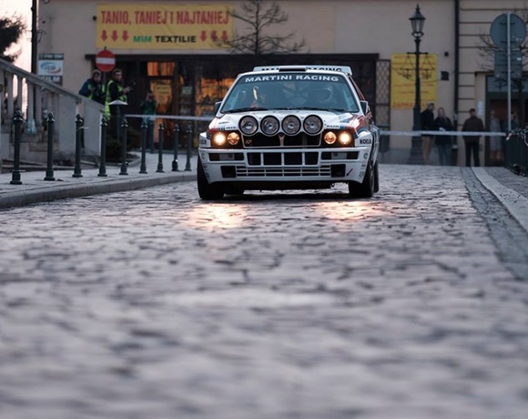 Rally car with Martini Racing livery and extra spotlights driving on a wet cobblestone street through a historic town, spectators nearby