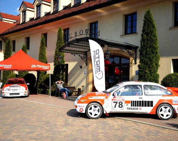 Hotel entrance with arched iron canopy, tiled roof and tall cypress shrubs; two race cars and branded tent displayed on paved forecourt.