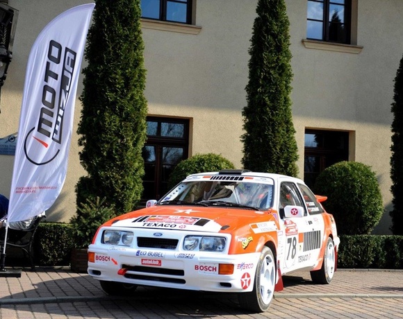 Orange-and-white rally car No. 78 parked on hotel courtyard by tall cypress trees and a sponsor flag