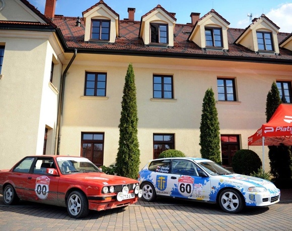 Historic inn facade with dormer windows, landscaped courtyard with tall cypress, rally cars and sponsor tent on cobblestone drive