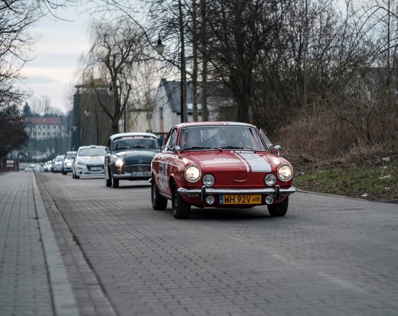Red vintage sports car leading a classic car rally along a tree-lined cobblestone street at dusk