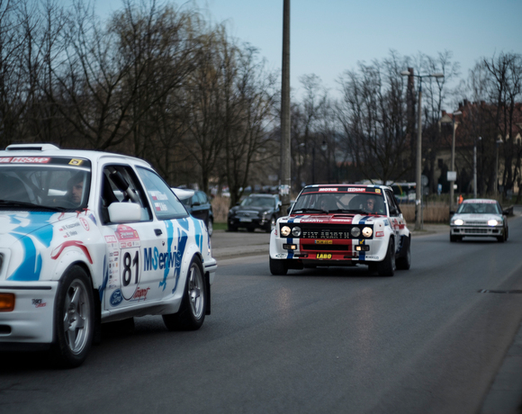 Three vintage rally cars with racing liveries driving on a town street under an overcast sky, drivers visible