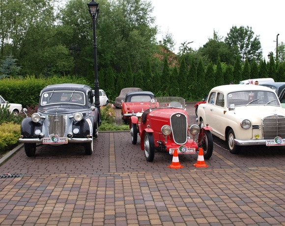 Three vintage cars — black sedan, red roadster with traffic cones, and cream saloon — parked on a paved driveway by a lamppost and hedge.