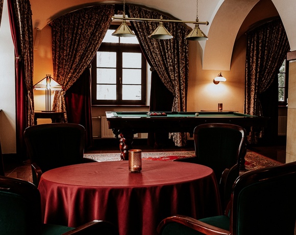 Elegantní hotelový salonek s kulečníkem, klenutými stropy, vzorovanými závěsy, teplým osvětlením a kulatým stolem s bordó ubrusem.