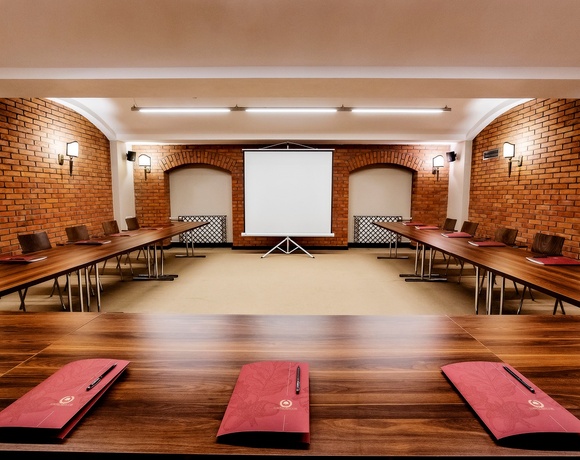 Konferenzraum mit U-förmigen Holztischen, Projektionsleinwand, freigelegten Ziegelwänden und warmen Wandlampen.