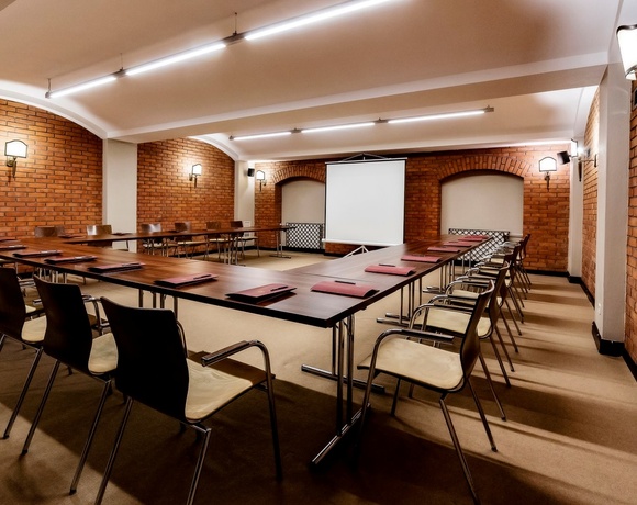 Konferenzraum mit Ziegelwänden, U-förmigen Tischen, Stühlen, Projektorleinwand und warmem Wandlicht