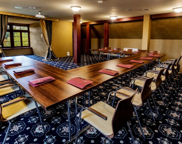 Konferenzraum in U-Form mit Holztischen, gepolsterten Stühlen, roten Ordnern an jedem Platz, Deckenprojektor und Fensterblick ins Grün.