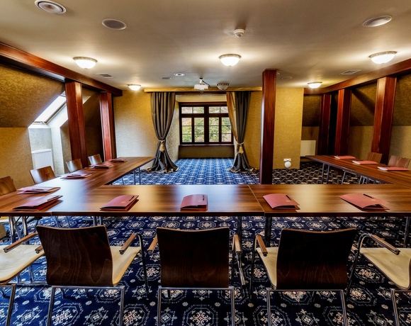 Hotel-Tagungsraum mit U-förmigen Holztischen, modernen Stühlen, gemustertem Teppich, Deckenprojektor, Dachfenster und Vorhängen