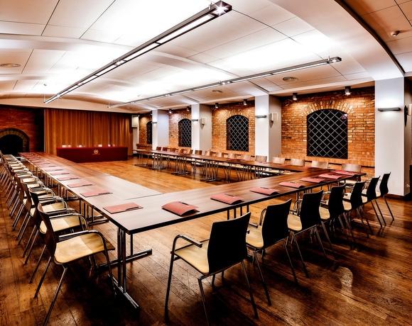 Konferenzraum in U‑Form: warmer Holzboden, freigelegte Backsteinbögen, lange Tische mit Notizblöcken, Deckenbeleuchtung, Wandlautsprecher und roter Rednertisch.