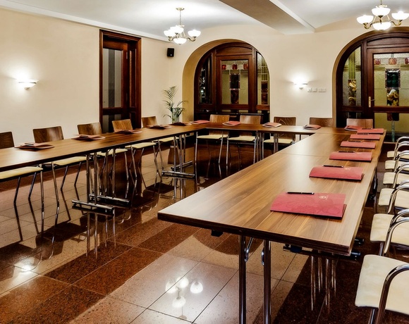 Konferenzraum in U‑Form mit Holztischen, gepolsterten Sesseln, roten Ordnern, Kronleuchtern und bogenförmigen Bleiglas-Türen bei warmer Beleuchtung