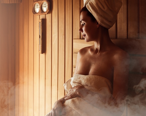 Woman wrapped in towel and turban relaxing in wooden hotel sauna with steam and wall thermometer