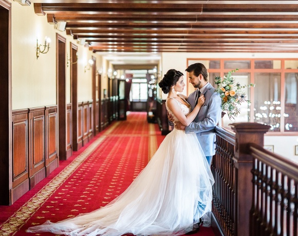 Bride and groom embracing on an elegant hotel balcony corridor with red carpet, wood paneling, chandeliers and bridal bouquet