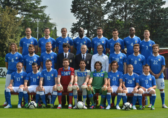 Men’s football team in blue kits and coaches posed on a sunlit pitch with soccer balls and goal, trees in the background