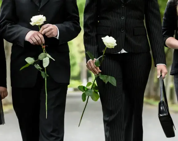 Two people in black formal attire holding white roses, walking on a tree-lined path