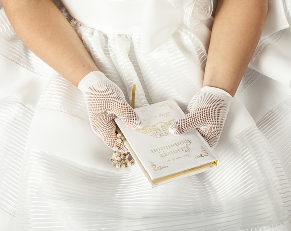 Child's gloved hands holding a white prayer book and pearl rosary on a layered white communion dress