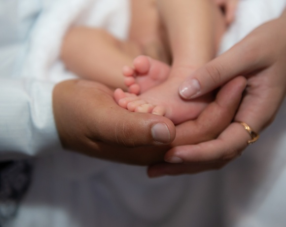Tender adult hands cradling a newborn's tiny feet on soft white linens, intimate family moment