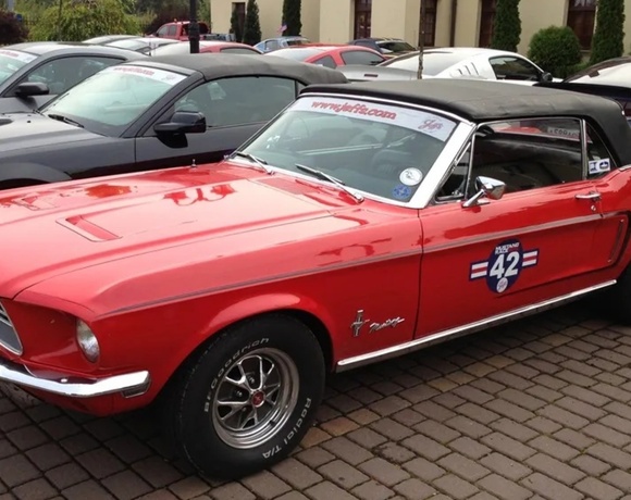 Red Ford Mustang convertible with black soft top and '42' race decal parked on cobblestone beside modern cars