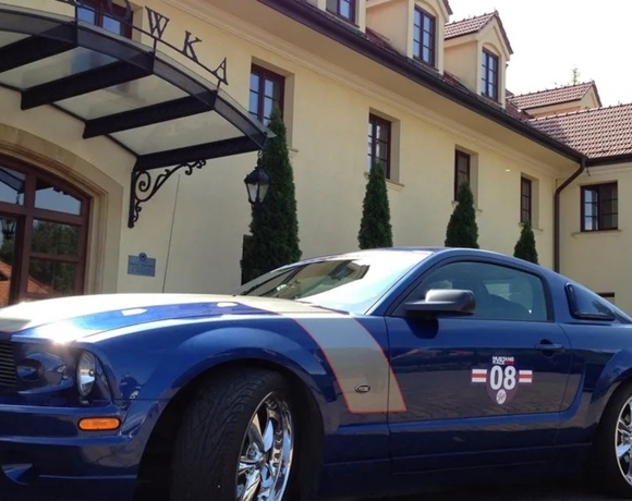 Elegant cream hotel entrance with glass-and-iron canopy, tall cypress shrubs and a blue sports car with chrome wheels parked outside.