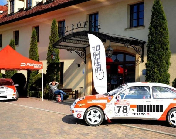 Hotel entrance with wrought-iron canopy, red-tiled roof and tall cypress trees; rally cars and branded tent create lively event display.