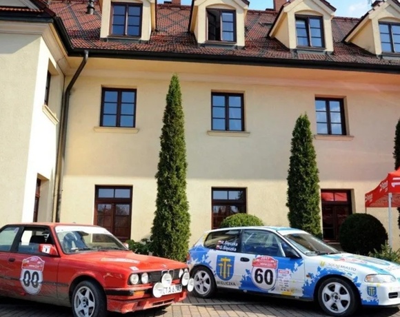 Cream historic hotel facade with tiled roof and dormer windows, courtyard parking with classic rally cars and red event tent