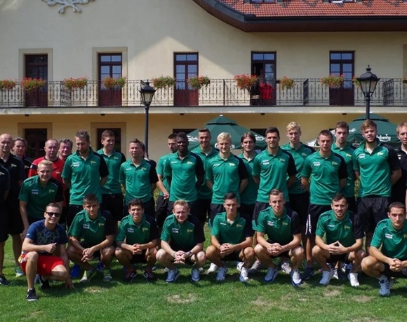 Men's sports team in green jerseys posed on hotel lawn in front of a two-story building with balcony flower boxes and umbrellas.