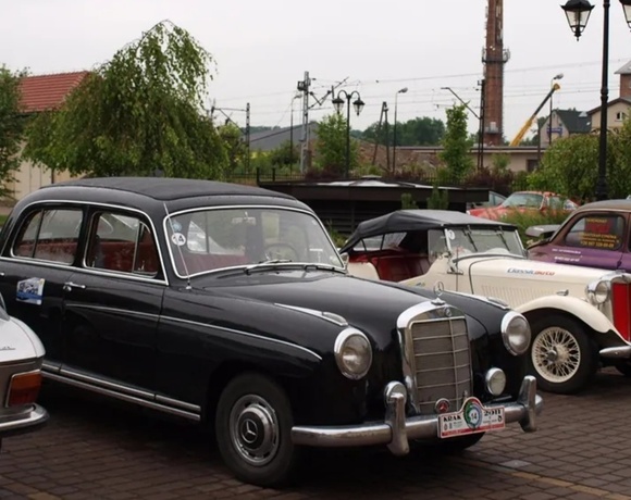 Row of classic vintage cars parked on a cobblestone courtyard with lamp posts and trees under an overcast sky