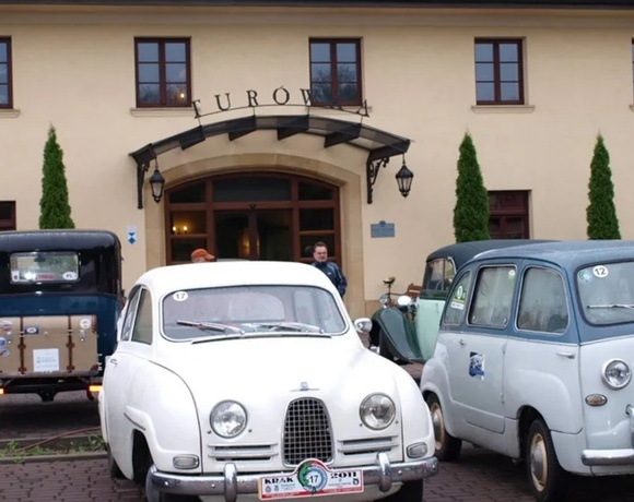 Beige hotel entrance with arched doorway, lanterns, tall cypress trees and vintage cars on a brick forecourt