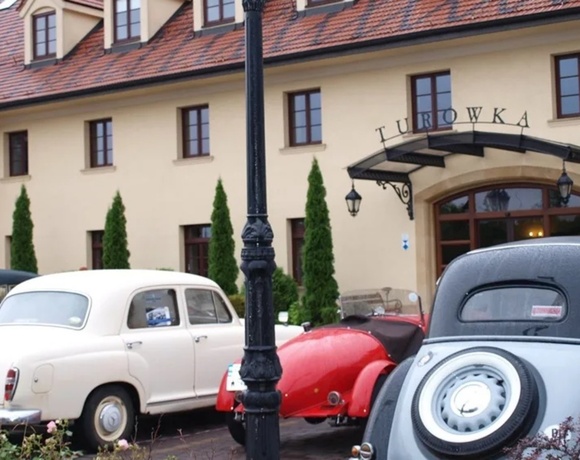 Historic-style hotel entrance with tiled roof, arched glass canopy, tall cypress shrubs and vintage cars parked in front