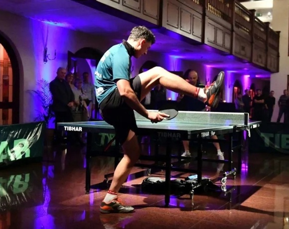 Acrobatic table-tennis show in hotel lobby with wooden balcony, purple uplighting, polished floor and watching guests.