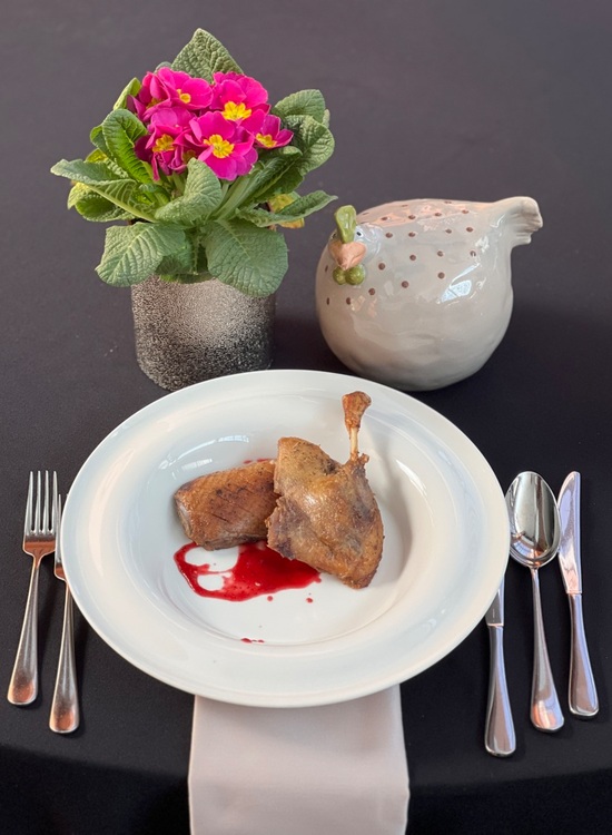 Udko kaczki z sosem z czerwonych owoców w białej misce, sztućce na czarnym obrusie, różowe kwiaty w doniczce i ceramiczny ptak