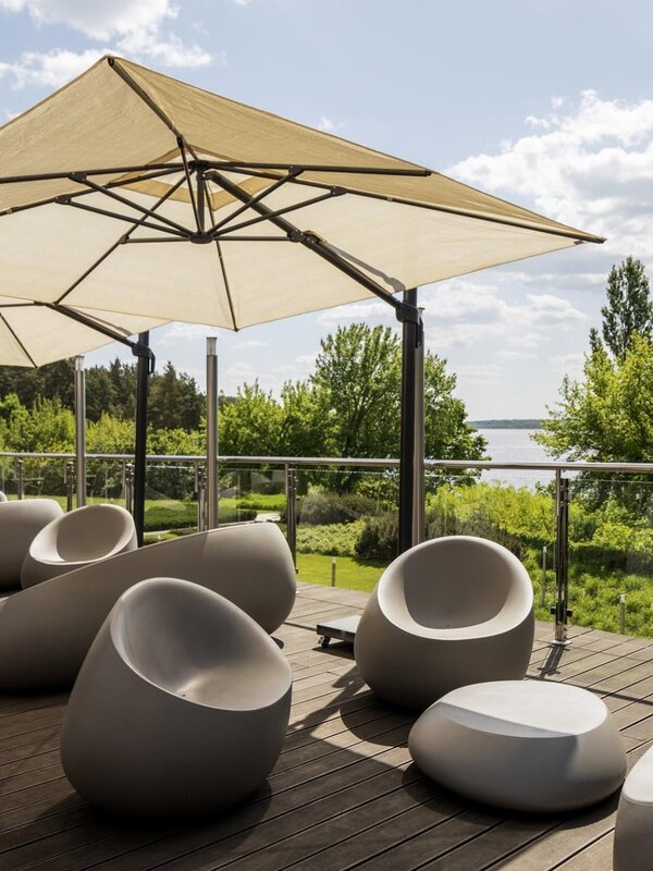 Sunlit hotel terrace with modern rounded lounge chairs, large parasols, glass railing and views over green gardens and a lake.
