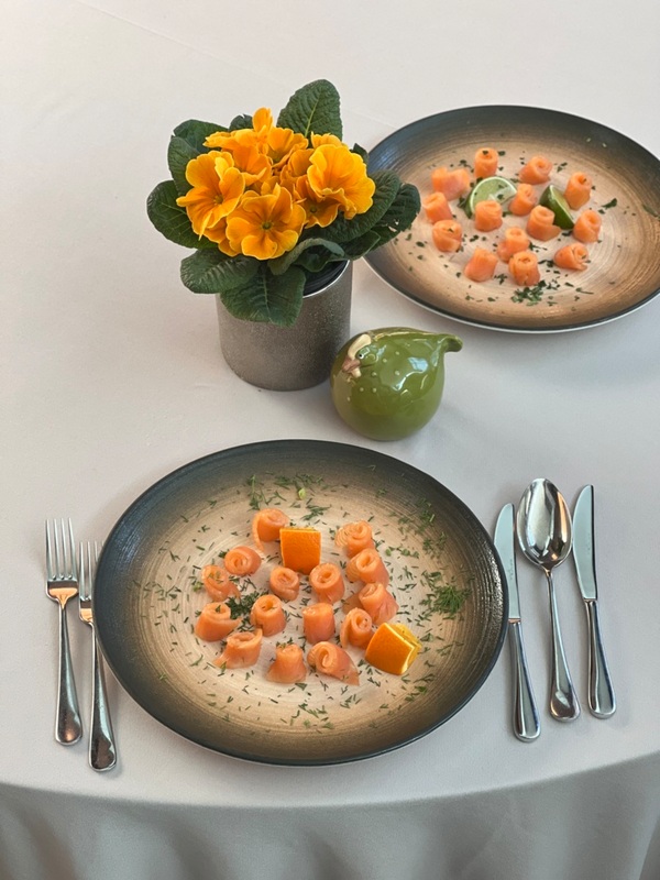 Stół dla dwojga z talerzami rozetek z łososia, cząstkami cytrusów i koperkiem oraz żółtą doniczką z kwiatami.