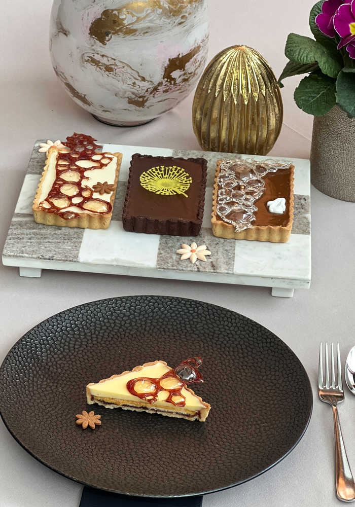 Trzy tarty na marmurowej tacy i kawałek cytrynowej tarty na ciemnym talerzu, złote wazy, pierwiosnek i srebrne sztućce