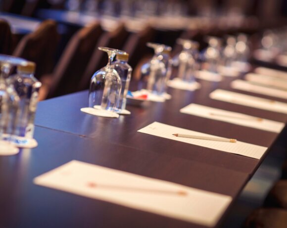 Sala konferencyjna z ustawionymi miejscami, kartkami, ołówkami i wodą na długim stole dla uczestników spotkania.