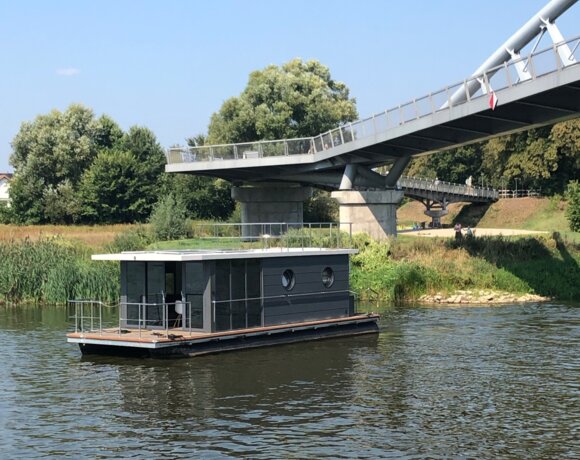 Nowoczesny, czarny domek na wodzie z okrągłymi oknami, na rzece przy zielonym brzegu i widocznym mostem spacerowym