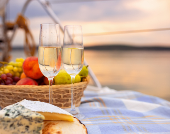 Dwa kieliszki szampana na kocu na tle kosza z owocami podczas romantycznego zachodu słońca nad wodą.