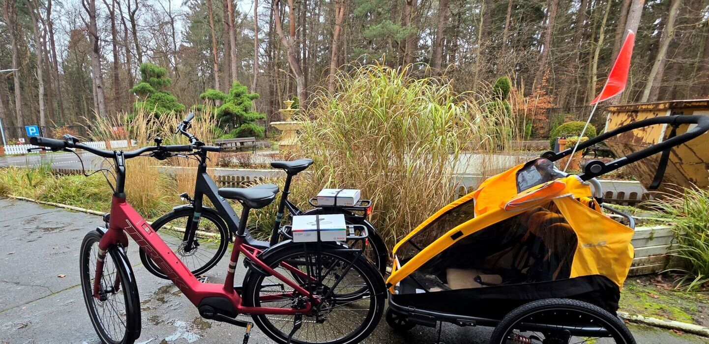 Dwa rowery elektryczne zaparkowane na ścieżce w zalesionym otoczeniu, jeden z nich z przyczepką dziecięcą w jasnopomarańczowym kolorze