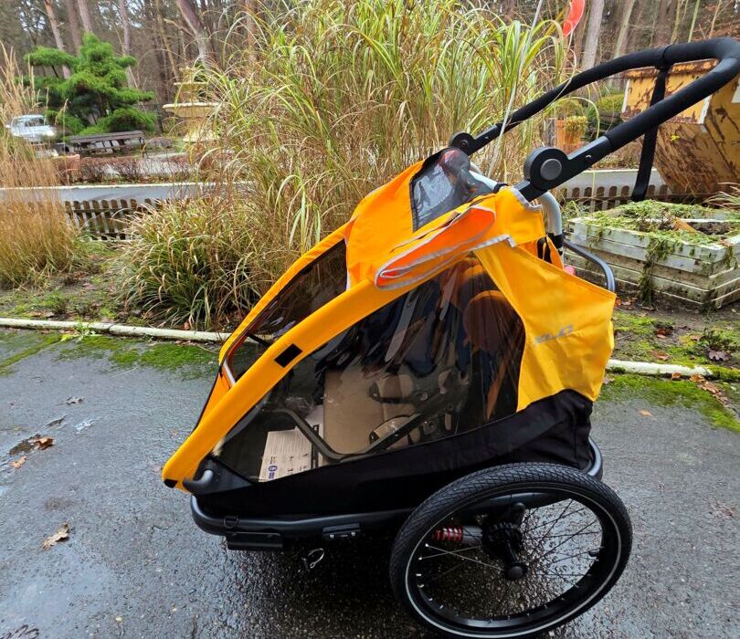 Pomarańczowy wózek dziecięcy z przezroczystą osłoną na tle leśnej ścieżki i jesiennych traw