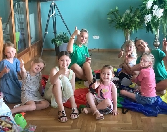 Kinder und zwei Betreuerinnen sitzen auf Parkett im Hotel-Kinderclub, bunte Spielmatten, Pflanzen, alle zeigen Daumen hoch.