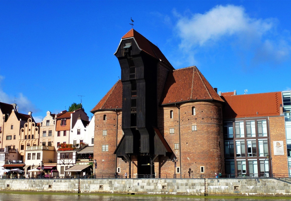 Medieval riverside crane and red‑tiled brick tower beside waterfront cafés with terraces offering river views