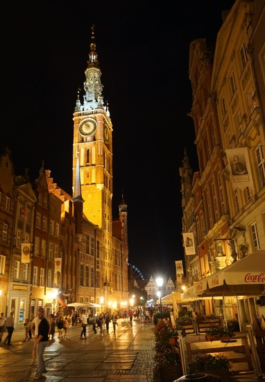 Historic clock tower and warm-lit pedestrian street with outdoor cafés and terraces, lively evening atmosphere