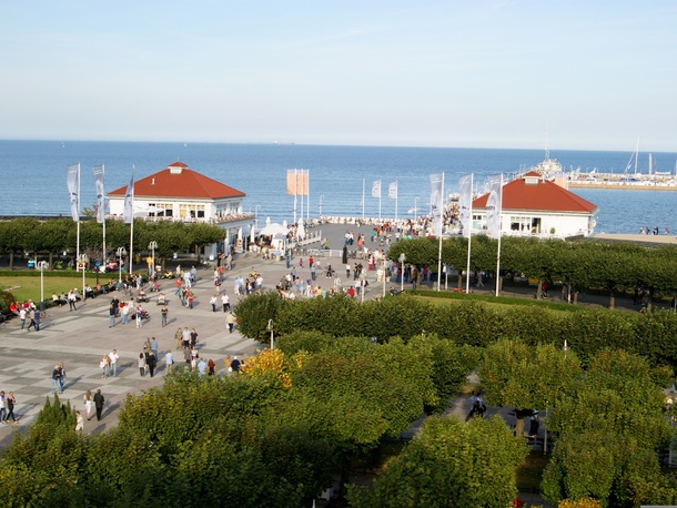 Nadmorska promenada z dwoma pawilonami o czerwonych dachach, tarasami restauracyjnymi, flagami i mariną, tłumy spacerujących przy morzu