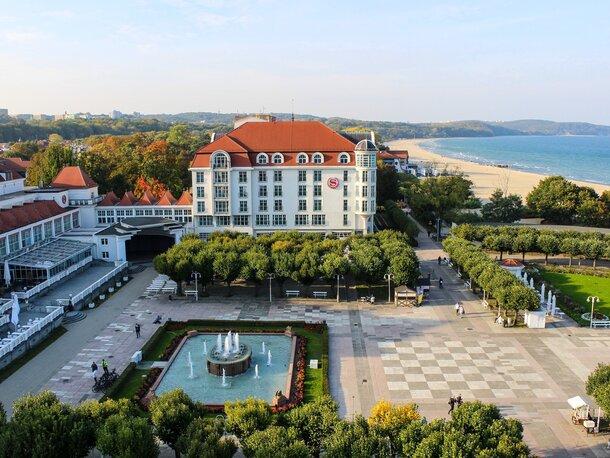 Hotel nadmorski z białą elewacją i czerwonym dachem, placem obsadzonym drzewami, fontanną, promenadą i piaszczystą plażą.