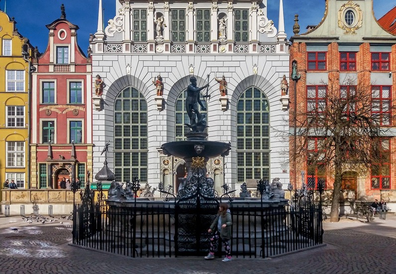 Ornate Neptune fountain before colorful gabled houses and a white Renaissance facade on a sunny square, passersby nearby.