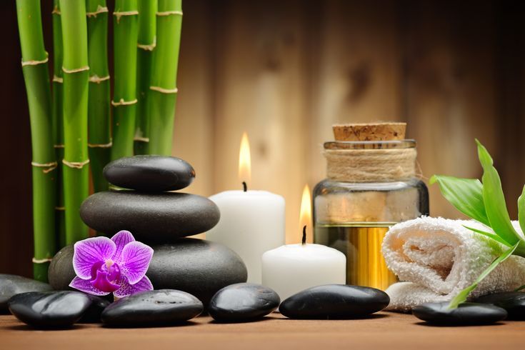 Raminanti SPA kompozicija: sukrauti juodi masažo akmenys, violetinė orchidėja, degančios žvakės, aliejus, susuktas rankšluostis ir bambukas.