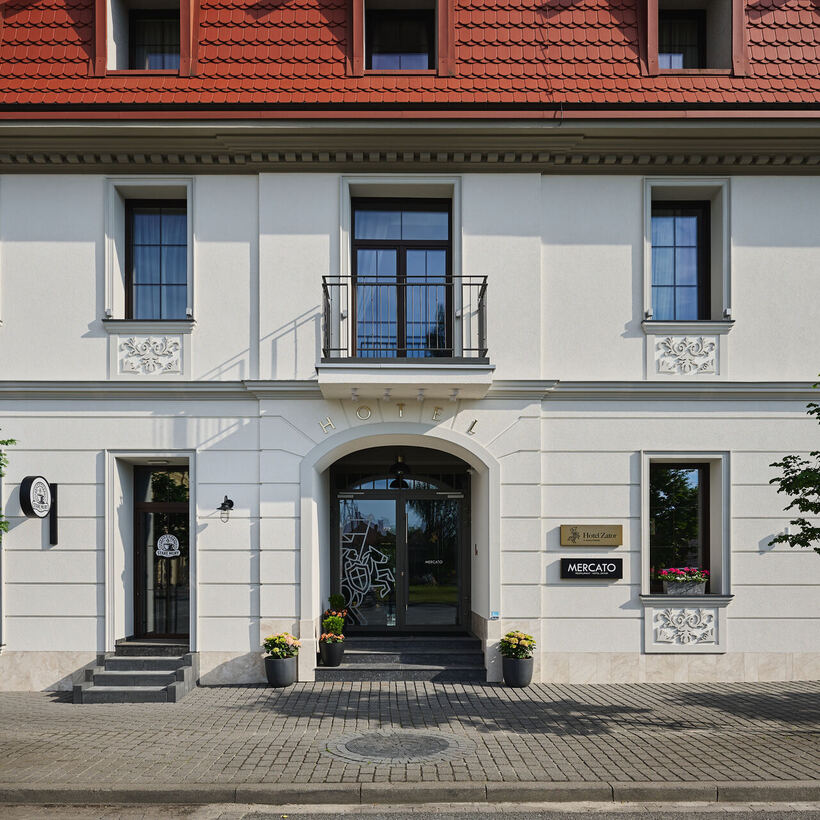 Elegante Fassade eines Boutique‑Hotels: weißer klassischer Frontbereich mit Bogenportal, Balkon, Blumenkästen und rotem Ziegeldach.