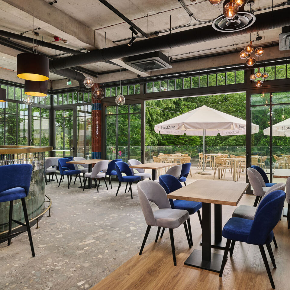 Jasna restauracja hotelowa ze ścianą okien, niebieskimi aksamitnymi krzesłami, łukowym kafelkowym barem, ciepłymi lampami i zielonym tarasem