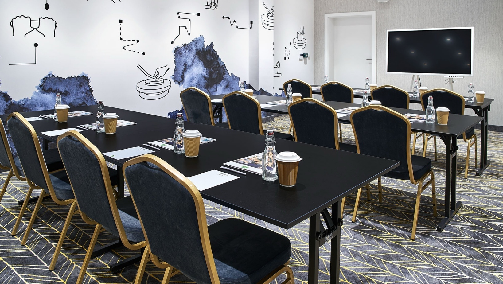 Sala konferencyjna hotelu w układzie szkoleniowym z czarnymi stołami, granatowymi krzesłami, ekranem, wodą i kawą.